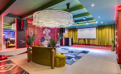 Арендовать Стильное пространство Астрид. Просторное помещение с зоной для отдыха, включает зеленый диван, проектор с экраном, декоративные растения, разноцветные ковры и плиточный пол. Потолочные светильники, красочные стены с крупным изображением человека и зелеными шторами.