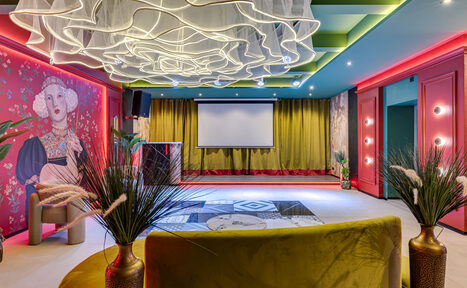 Арендовать Стильное пространство Астрид. Просторное помещение с диванами, проектором и экраном. Яркие стены украшены художественными рисунками. Потолок с декоративным светильником. Пол с ковром в геометрический узор. Зеленые и розовые акценты в интерьере. Растения в горшках.