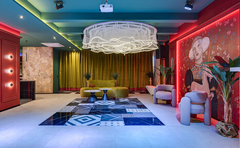 Арендовать Стильное пространство Астрид. Просторная комната с высокими потолками и цветной плиткой на полу. В центре висячий декоративный светильник. По периметру разноцветные стены с занавесками и крупным изображением. Мягкая мебель: кресла, диван. Растения в горшках.
