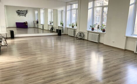 Арендовать Территория Танца. Просторное помещение с деревянным полом и большими окнами. На одной стене большое зеркало. Вдоль окон размещены растения, радиаторы и вентиляторы. В углу фиолетовый диван. На стене текст. Светлые стены и потолок.