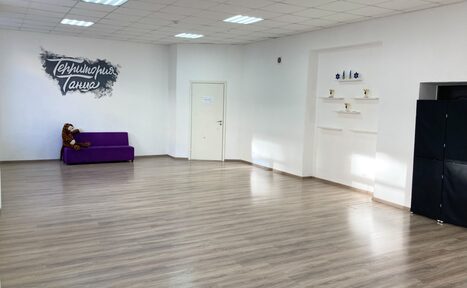 Арендовать Территория Танца. Просторная светлая комната с ламинатным напольным покрытием и белыми стенами. В углу фиолетовый диван и мягкая игрушка. На стене граффити с надписью "Территория танца". Потолок с квадратными светильниками, полки с декором.