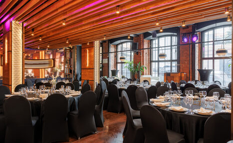 Арендовать Zafferano Loft(Зафферано лофт). Пространство с деревянным потолком и кирпичными стенами, круглыми столами, сервированными стеклянной и столовой посудой, черными чехлами на стульях. Большие окна, точечное освещение, подвесные светильники, зеленые растения, полки с книгами.