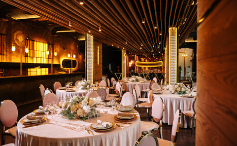 Арендовать Zafferano Loft(Зафферано лофт). Банкетный зал с круглой расстановкой столов, покрытых белыми скатертями и украшенных цветочными композициями. Розовые мягкие стулья с металлическими ножками. Потолок с деревянными балками и точечными светильниками. Стены кирпичные, теплое освещение.