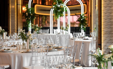 Арендовать Zafferano Loft(Зафферано лофт). Банкетный зал с круглыми столами, накрытыми белыми скатертями, и прозрачными стульями. На столах бокалы и белые цветы. Арки с неоновыми огнями и зеленью. Лёгкое декоративное освещение, стены с золотыми и белыми элементами.