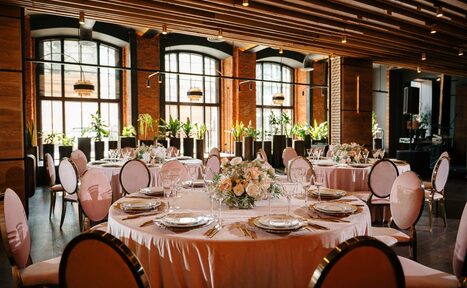 Арендовать Zafferano Loft(Зафферано лофт). Большой зал с круглыми столами, покрытыми белыми скатертями и сервированными тарелками и бокалами. Овальные стулья с белыми спинками. Высокие окна с черными рамами. Растения на подоконниках. Потолок с деревянными панелями и подвесными светильниками.