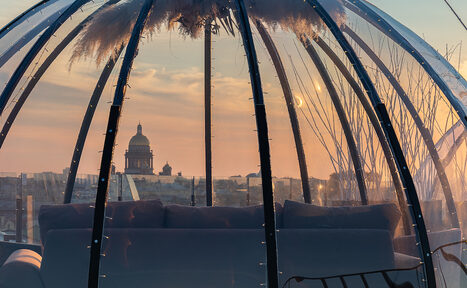 Арендовать Sky Terrasse(Небо Терраса). Прозрачный купол с панорамным видом на город, конструкция из металлических дуг. Внутри темный диван, светодиодная подсветка, подвесной декор. На горизонте городской пейзаж с куполом большого здания, закатное освещение.