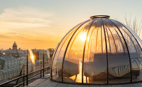 Арендовать Sky Terrasse(Небо Терраса). Купольная стеклянная конструкция на крыше с видом на город. Внутри находятся диваны. Прозрачные стены отражают солнечный свет. Пол выложен плиткой. Пространство окружено стеклянными ограждениями. Закатное солнце на горизонте.