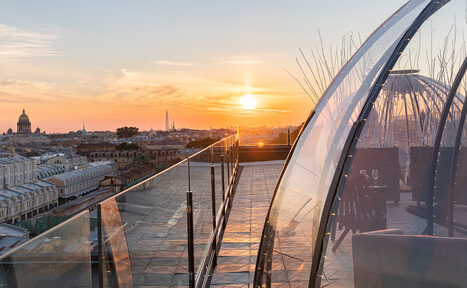 Арендовать Sky Terrasse(Небо Терраса). Открытая терраса на крыше с прозрачными куполами, стеклянными перилами и плиточным полом. Несколько столов и стульев внутри купола. Панорама города на фоне заката.