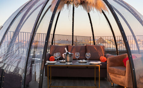 Арендовать Sky Terrasse(Небо Терраса). Современный купол из прозрачного материала на крыше, внутри коричневый диван и кресло, низкий стол с белыми тарелками, бокалами и ведёрком с бутылкой, украшение из сухих растений сверху, вокруг металлическое ограждение, вечернее освещение.