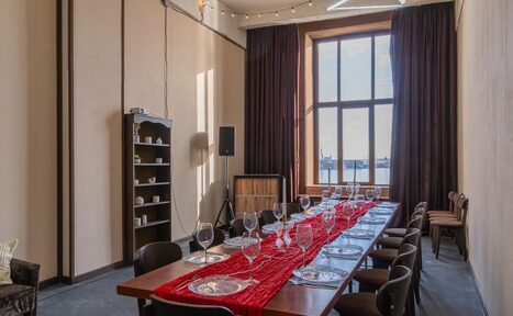 Арендовать Chocolate Loft (Шоколад Лофт). Зал с видом на Финский залив. Прямоугольное столовое пространство с длинным деревянным столом, накрытым красной скатертью и сервировкой на 12 персон. Металлические стулья, высокий стеллаж с посудой, светлые стены, большая штора, большое окно, черные портьеры, струнные светильники.