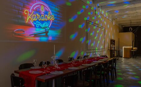 Арендовать Chocolate Loft (Шоколад Лофт). Зал с видом на Финский залив. Просторное помещение с длинным деревянным столом, покрытым красной дорожкой, сервировано бокалами и свечами. На стене неоновая надпись "Karaoke" и микрофон. Тёмные стулья, разноцветное освещение, белый кирпич, барная стойка в глубине.