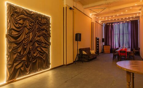 Арендовать Chocolate Loft (Шоколад Лофт). Зал с видом на Финский залив. Лофт-пространство с высоким потолком, обеденным столом и стульями. Тёмные шторы на окнах, драпировка на стене с подсветкой, диван, акустические колонки. Потолочное освещение из гирлянд, пол тёмно-серый, стены бежевые.