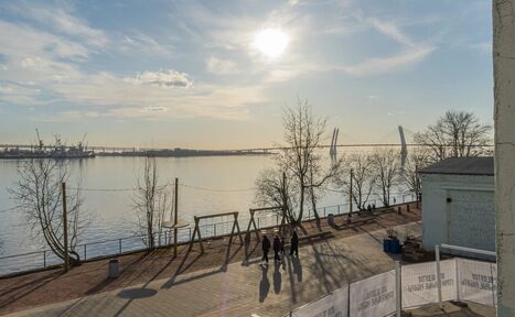 Арендовать Chocolate Loft (Шоколад Лофт). Зал с видом на Финский залив. Променад вдоль реки с асфальтированной дорожкой, несколько деревьев без листвы, лавочки, находящиеся на солнце, вдалеке мост, спокойная вода, небо с облаками, группа людей, ограждение с надписями, металлические конструкции.