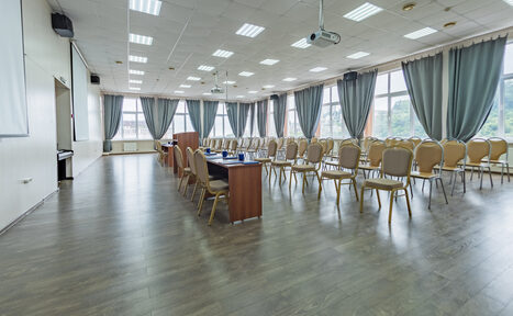 Арендовать Конференц зал в отеле Гарден Хиллс 3. Большая комната с деревянным полом, кафельным потолком с встраиваемыми светильниками, длинными рядами стульев и деревянными столами. Большие окна с серыми шторами, проекторы, экраны и кондиционеры на стенах.