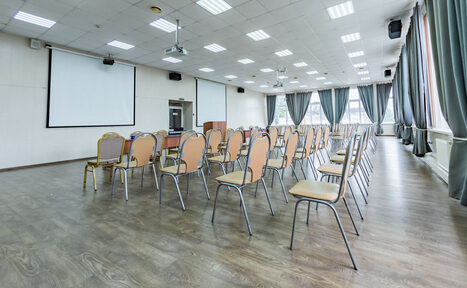 Арендовать Конференц зал в отеле Гарден Хиллс 3. Конференц-зал с деревянным полом, белыми стенами и подвесным потолком. Ряды стульев, проекционный экран, проекторы, аудиоколонки, большие окна с серыми шторами. Посредине зала установлена кафедра.