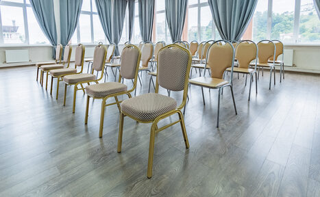 Арендовать Конференц зал в отеле Гарден Хиллс 3. Просторное помещение с рядами стульев бежевого цвета. Пол из светлого дерева, большие окна с серыми шторами. Светлые стены, естественное освещение из окон. Стулья расположены в несколько рядов.