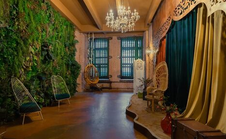 Арендовать Polygon(Полигон). Зал Марокко. Пространство с зелёной стеной из растений, двумя креслами-коконами, качелями, сценой с драпировкой и деревянным декором. Потолок с открытыми балками, люстра и настенные светильники. Цвета: зелёный, бежевый, коричневый. Пол бетонный, стены из кирпича.