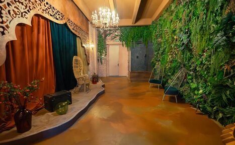 Арендовать Polygon(Полигон). Зал Марокко. Пространство с деревянным полом коричневого цвета, на стенах зелёные растения. Справа два стула с металлическими спинками, слева плетёное кресло, драпировки оранжевого и тёмно-зелёного цвета. Декоративная люстра, деревянный декор на стене.