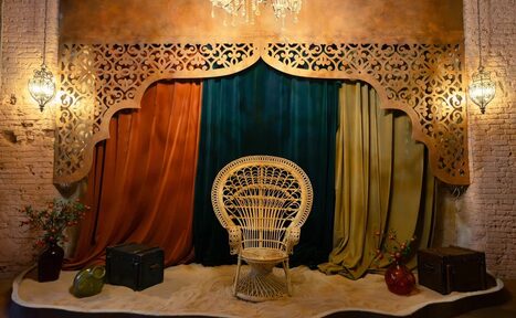 Арендовать Polygon(Полигон). Зал Марокко. Декорированное пространство с узорной аркой, драпированное тканями оранжевого, синего и зелёного цветов. Плетёное кресло в центре. По бокам коричневые чемоданы и вазы с цветами. Стены из светлого кирпича. Подвесные фонари и декоративная люстра.