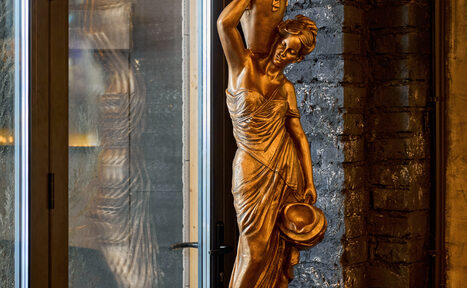 Арендовать Tangiers Lounge(Танжер Лаундж) . Статуя женщины из бронзового материала рядом с кирпичной стеной и стеклянной дверью. Женщина держит кувшин над головой и шляпу в другой руке. Теплое освещение.