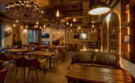 Арендовать Tangiers Lounge(Танжер Лаундж) . Зал с деревянными столами и кожаными стульями. Потолок украшен люстрами и лампами с теплым освещением. Стены декорированы кирпичом и деревом, есть большие оконные проемы. На стенах полки с книгами и картинами. Теплые коричневые и бежевые тона.