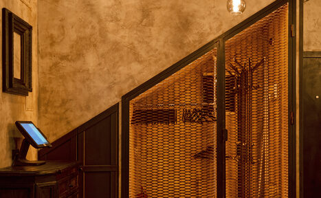 Арендовать Tangiers Lounge(Танжер Лаундж) . Коридор с индустриальными элементами, бежевые стены с текстурой, металлическая сетчатая перегородка, деревянная мебель с монитором, настенный крючок, подвесная лампа с теплым светом, черные и коричневые оттенки, строгая геометрия.