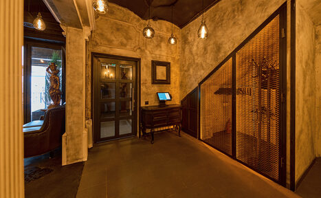 Арендовать Tangiers Lounge(Танжер Лаундж) . Интерьерное пространство с коричневыми стенами в стиле лофт, стеклянная дверь, низкое освещение с подвесными лампами, деревянная мебель, металлическая перегородка, пол темный, присутствует оборудование на столе, зеркальная колонна.