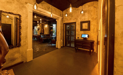 Арендовать Tangiers Lounge(Танжер Лаундж) . Коридор с арочным входом в зал. На стенах декоративная штукатурка. Подвесные лампы создают тёплое освещение. Справа стол с монитором. Слева зеркало. За аркой видны столы и кресла в тёмных тонах. Потолок тёмный.