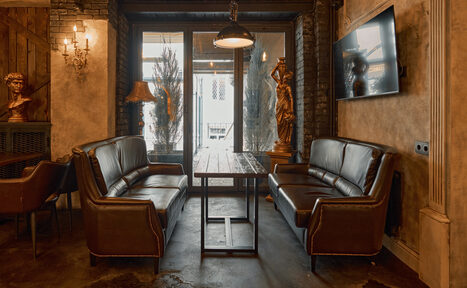 Арендовать Tangiers Lounge(Танжер Лаундж) . Пространство с двумя коричневыми кожаными диванами и деревянным столом между ними, на стене телевизор, рядом деревянная статуя, подвесной светильник и бра, окна с декоративными растениями, кирпичные стены и полированный бетонный пол.