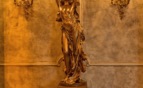 Арендовать Tangiers Lounge(Танжер Лаундж) . Золотая статуя в классическом стиле с изображением женщины стоит на постаменте перед текстурированной стеной теплого коричневого оттенка. Справа и слева позолоченные декоративные элементы с орнаментом. Освещение теплое.