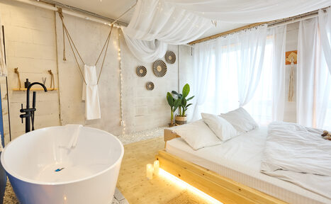 Арендовать Пляжный лофт с галькой, открытой ванной, панорамными окнами. Спальня со светлыми стенами и потолком с белым декором, большая кровать с белым постельным бельём, отдельно стоящая белая ванна, чёрный смеситель, декоративные тарелки на стене, зелёное растение в горшке, свечи, деревянный пол.