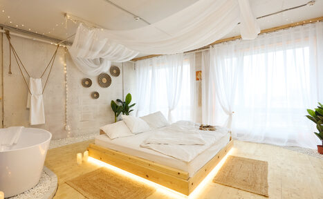 Арендовать Пляжный лофт с галькой, открытой ванной, панорамными окнами. Пространство с деревянным полом и светлыми стенами. В центре кровать на подиуме с белыми покрывалами и подушками. Справа ванна округлой формы, рядом дорожки из ковров. Большие окна с белыми занавесками. Украшение на стене и несколько горшков с растениями.