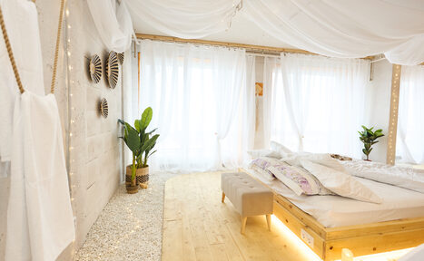 Арендовать Пляжный лофт с галькой, открытой ванной, панорамными окнами. Светлая спальня с кроватью на деревянном подиуме, фоновое освещение. Белые занавески на окнах, растения в горшках, серая скамья у изножья кровати. Стены украшают декоративные элементы, пол из светлого дерева и белых камней.