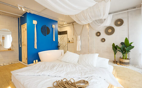 Арендовать Пляжный лофт с галькой, открытой ванной, панорамными окнами. Спальня с большой кроватью и белым постельным бельём. Белые стены и деревянный пол, стены украшены декором. Ванна в комнате возле ярко-синей стены. Зелёные растения в углу. Белые тканевые драпировки на потолке.