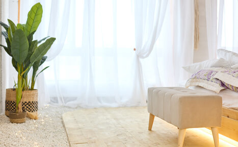 Арендовать Пляжный лофт с галькой, открытой ванной, панорамными окнами. Светлая комната с деревянным полом и белыми прозрачными шторами. Мягкая скамья с бежевой обивкой у изножья кровати. По бокам зелёные растения в плетёных корзинах. Светлое постельное бельё, подушки с узором сиреневых оттенков.