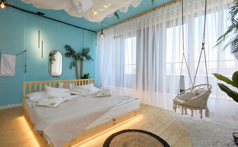 Арендовать Лофт Мальдивы с открытым душем, балконом и видом на Сити. Спальня с голубыми стенами, кровать с деревянным каркасом, подвесное кресло, пол из светлого дерева, прозрачные шторы, декоративные растения, подвесные лампы, овальное зеркало, белый текстиль на кровати, плетеный ковер.