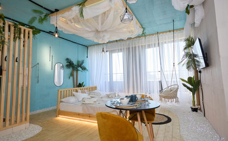 Арендовать Лофт Мальдивы с открытым душем, балконом и видом на Сити. Спальня с бирюзовыми стенами, кроватью под балдахином, деревянным полом. Обеденный стол с желтыми стульями, балкон с прозрачными шторами. Подвесное кресло, множество растений, телевизор на стене. Декоративные элементы из ткани и геометрические светильники.