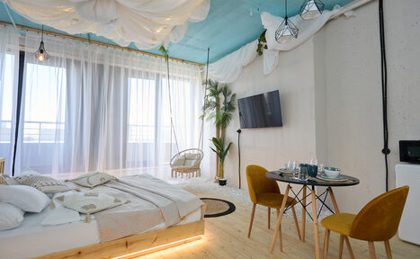 Арендовать Лофт Мальдивы с открытым душем, балконом и видом на Сити. Спальня с деревянным полом, низкой кроватью, двумя желтыми стульями и круглым обеденным столом. Настенный телевизор, подвесное освещение, белые занавески на окнах, голубой потолок с драпировкой. Декоративные растения и подвесное кресло.