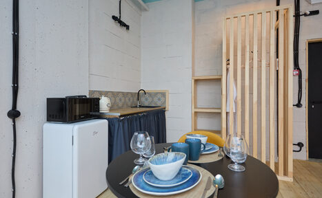 Арендовать Лофт Мальдивы с открытым душем, балконом и видом на Сити. Маленькая кухонная зона со столом и двумя стульями. На столе три тарелки, чашки и бокалы. В углу мини-холодильник и микроволновка. Раковина с узорчатым фартуком. Слева от раковины текстильная штора. Светлые стены и деревянная перегородка.
