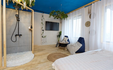 Арендовать Лофт Бали с фото зоной на 23 этаже с видом на Москва-реку. Комната с синем потолком и деревянным полом. Открытая душевая с черной сантехникой. Белые шторы на окнах, кресло-мешок, небольшой стол, подвешенные растения. На стене телевизор, декор в виде зелени и тарелок. Светлое покрывало на кровати.