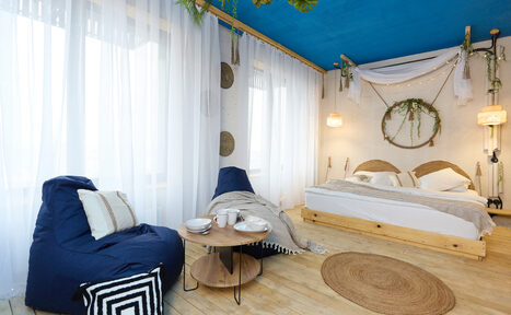 Арендовать Лофт Бали с фото зоной на 23 этаже с видом на Москва-реку. Спальня с двухместной кроватью, двумя подвесными светильниками, круглым деревянным столом и синим креслом-мешком. Белые стены и занавески, синий потолок, деревянный пол. Декор из текстиля и растений, круглое зеркало в изголовье кровати.