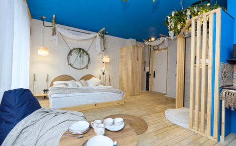 Арендовать Лофт Бали с фото зоной на 23 этаже с видом на Москва-реку. Просторная комната с синим потолком, деревянным полом и светлыми стенами. Двуспальная кровать с плетеными изголовьями. Деревянный стол с посудой, подвесные светильники, зеленые растения, диван, белый текстиль, деревянные перегородки, декоративные элементы на стенах.