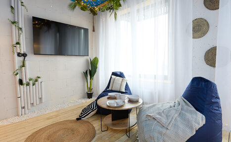 Арендовать Лофт Бали с фото зоной на 23 этаже с видом на Москва-реку. Комната с деревянным полом и белыми кирпичными стенами, телевизор на стене, два мягких кресла-мешка, круглые столики с посудой, большое окно с прозрачными шторами, плетёные декоративные элементы на стене, зеленые растения, светлое и уютное освещение.