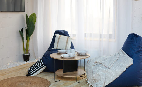 Арендовать Лофт Бали с фото зоной на 23 этаже с видом на Москва-реку. Комната с деревянным полом, двумя синими креслами-мешками, круглым деревянным столиком, несколькими чашками, пледами и подушками. Белые прозрачные занавески, комнатное растение в углу, настенное освещение.