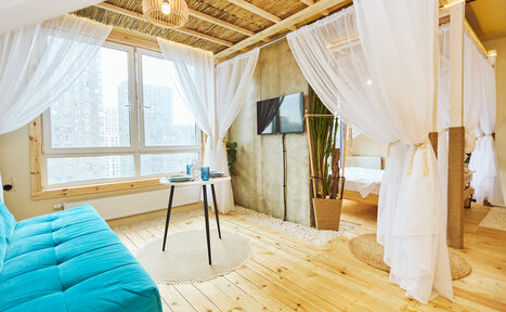 Арендовать Тропический лофт с бунгало, баром и французским балконом. Просторное светлое помещение с деревянным полом и потолком. Большое окно, разделённое занавесками на зоны. Бирюзовый диван, круглый стол с двумя стаканами, телевизор на стене, крупное растение в горшке. Светлая ткань занавесок, светильник на потолке.