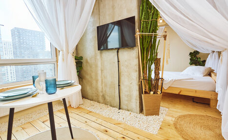 Арендовать Тропический лофт с бунгало, баром и французским балконом. Просторная комната с деревянным полом и бетонной стеной. Круглый белый стол с двумя приборами, телевизор на стене, кровать с белым покрывалом. Растения в горшке, белые занавески, мягкое дневное освещение, большое окно, коврик из натурального материала.