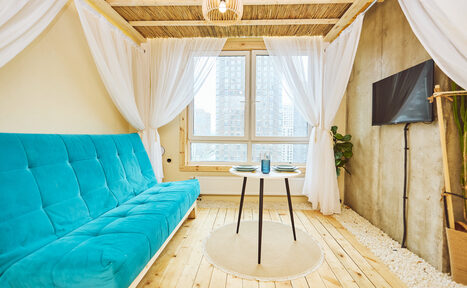 Арендовать Тропический лофт с бунгало, баром и французским балконом. Пространство с голубым диваном, круглым столиком с черными ножками, большая окно с белыми занавесками, деревянный пол, бетонная стена, небольшой ковер. Стена украшена растением и телевизором. Естественное освещение.