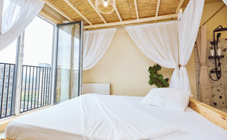 Арендовать Тропический лофт с бунгало, баром и французским балконом. Спальня с двуспальной кроватью, белыми простынями и подушками. Над кроватью белые полупрозрачные шторы. Открытые двери на балкон с черными металлическими перилами. Пол светлый, стены бежевые. Потолок из дерева с декоративным светильником.