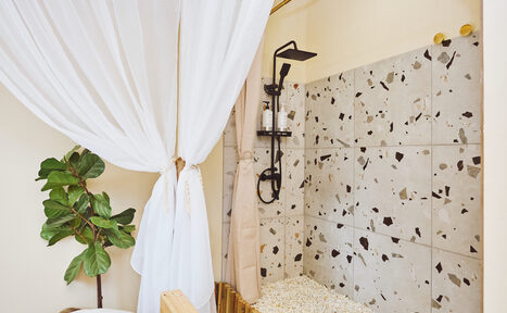 Арендовать Тропический лофт с бунгало, баром и французским балконом. Душевая зона с терраццо плиткой, черный душевой комплект, прозрачные диспенсеры для шампуня. Светлый текстильный занавес, пастельные стены. Зеленое растение в углу. Пастельные и нейтральные тона.