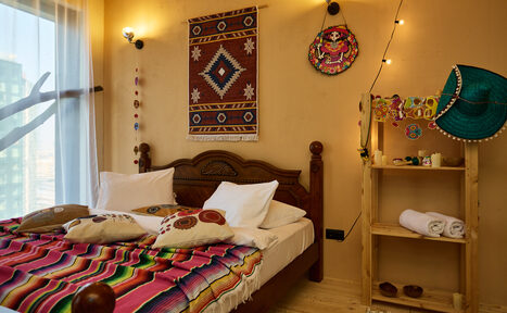 Арендовать ⁠Лофт в Мексиканском стиле для костюмированных вечеринок. Спальня с деревянной кроватью, покрытой разноцветным пледом и подушками. Стена украшена текстильным панно и декоративными тарелками. Рядом деревянный стеллаж с полотенцами. Стены светло-коричневые, на полу светлая древесина. Естественное и точечное освещение.